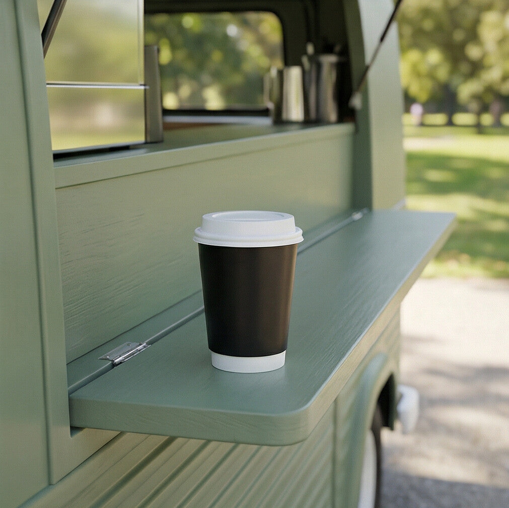 Black 12oz double wall paper cup with a white lid on the fold-down shelf of a sage green coffee van, park in background