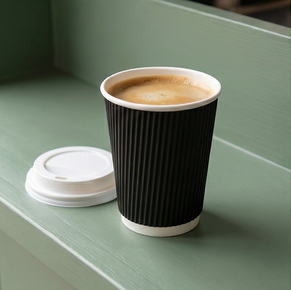 12oz Black Ripple Wall Paper Cup with frothy coffee inside, white lid beside on a sage green café shelf
