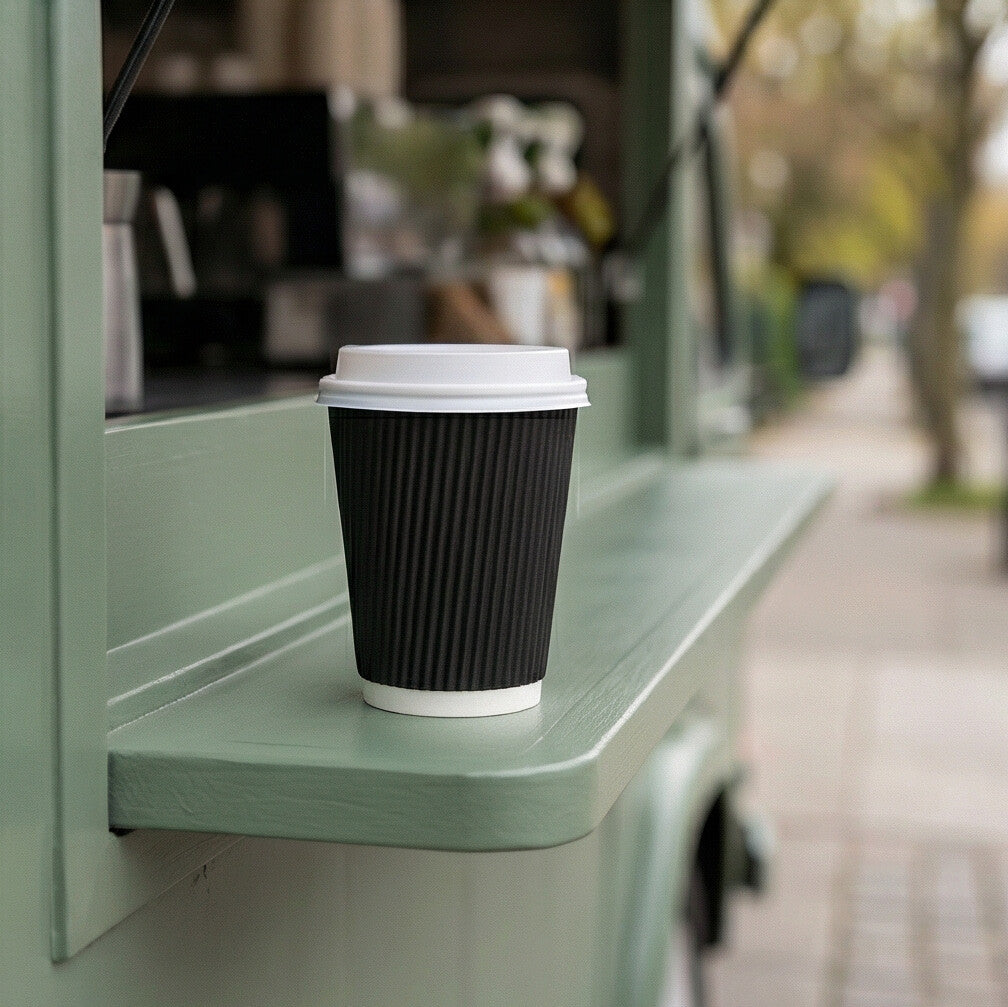 12oz Black Ripple Wall Paper Cup with a white lid on a sage green coffee van shelf, street and trees visible behind
