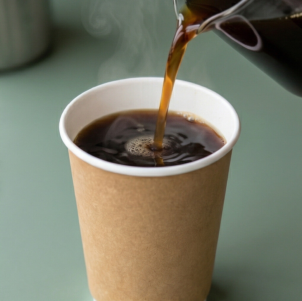Black coffee being poured from a glass jug into a 16oz kraft double wall paper cup on a sage green counter, steam rising
