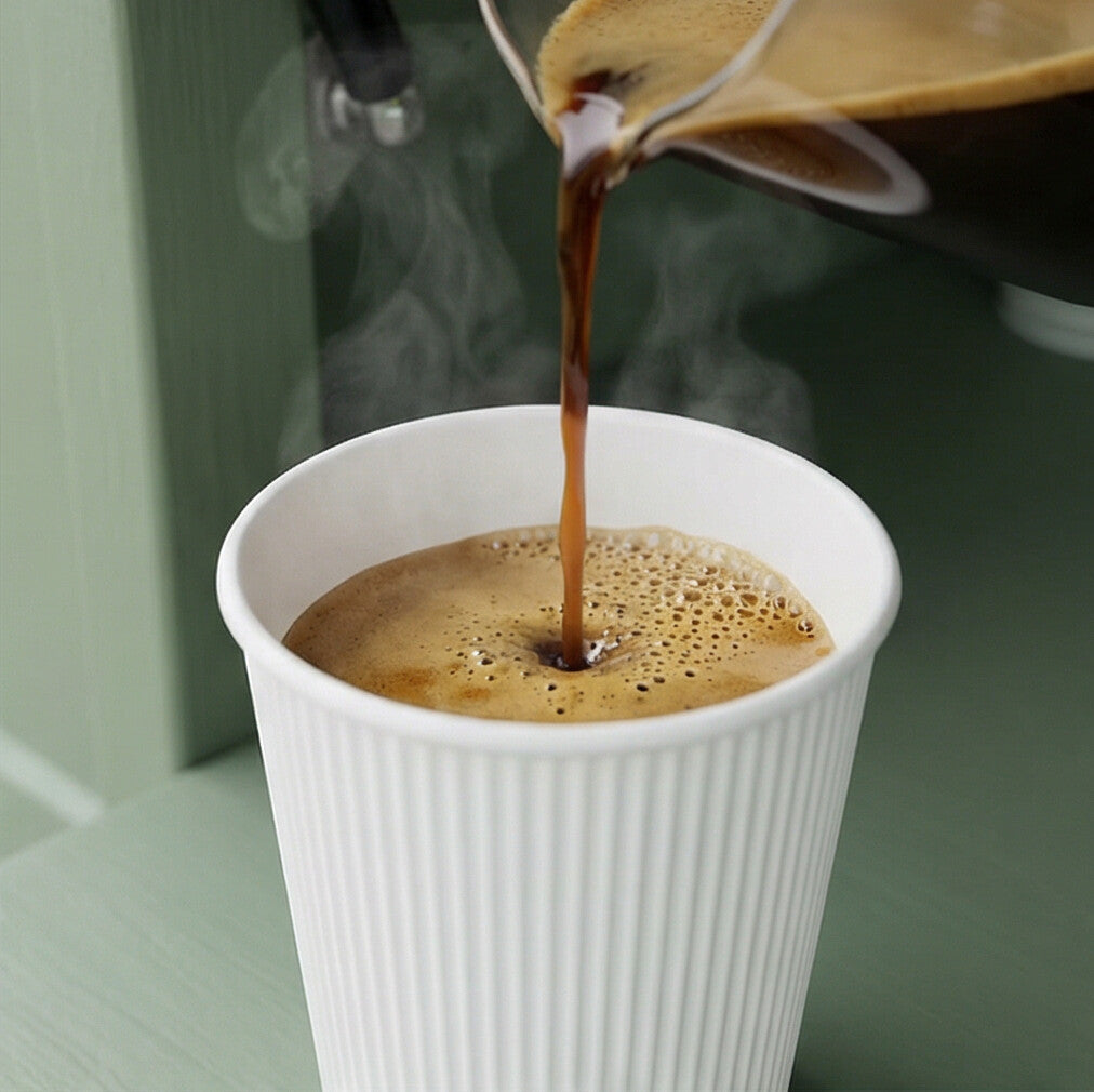 Hot coffee poured from a jug into an 8oz White Ripple Wall Paper Cup, steam rising, sage green café background