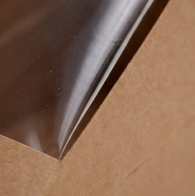 Kraft sandwich wedge deep fill box, extreme close-up detail of clear plastic window edge where plastic film meets natural kraft brown board construction on white background